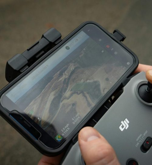 Hands controlling a drone with a smartphone display showing aerial view, demonstrating modern technology use.