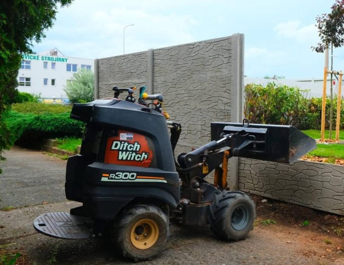 Nakladač Ditch witch R300 stojí na cestě u plotu